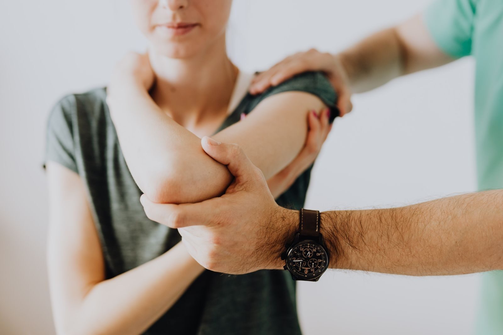 Crop faceless osteopath in modern wristwatch and uniform bending arm of anonymous thoughtful woman in casual wear in doctor office in hospital